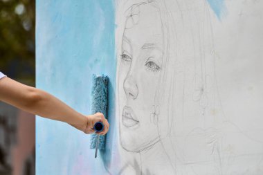 El, beyaz duvardaki genç kadın yüzünün çiziminin üzerine yumuşak mavi boya sürüyor. Doğal dış mekan, hafif ışıklandırma, sanatsal gelişim