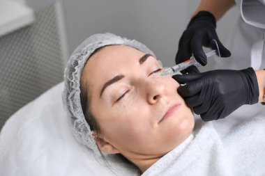 Kadın hasta klinik çevrede profesyonel olarak kozmetik enjeksiyondan geçiyor. Ayarlama sakin ve sterildir, estetik tedavilerde hassasiyet ve özen vurgulanır