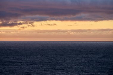Gün batımında sersemletici bir okyanus manzarası. Canlı turuncu ve pembe bulutlar durgun mavi su ve dramatik gökyüzüne karşı.