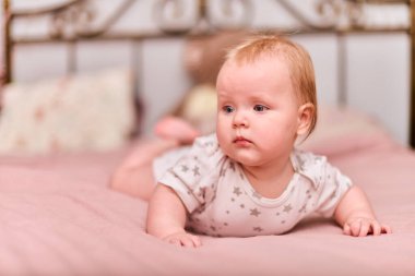 Açık renk saçlı, açık tenli, pembe yatak örtüsünde mavi gözlü bir bebek. Yıldız desenli tulum bezi. Ilık ışık rahat ve nazik bir atmosfer yaratır.