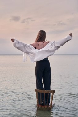 Genç bir kadın, sakin sularda tahta sandalyenin üzerinde sevinçle kollarını uzatır. Sahne, gökyüzü altında özgürlük ve sükunet hissi uyandırıyor. Bu da doğayla derin bir bağ olduğunu gösteriyor.