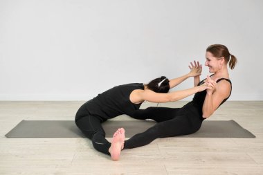Minimalist stüdyoda iki yetişkin kadın yoga pozu veriyor. Biri öne eğiliyor, diğeri onu destekliyor, bağlantı ve denge hissi yaratıyor.