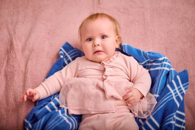 Küçük kız, çocuk, sarı saç, beyaz, pembe elbise giymiş, yumuşak, mavi kareli battaniyeye uzanmış. Konforlu ortam, sıcak aydınlatma, gevşek ifade, kapalı ortam