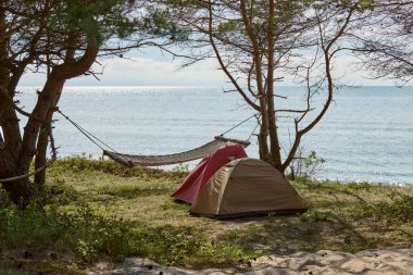 Huzurlu sahildeki ağaçların altına iki çadır ve hamak kurulur. Sahne, rahatlamayı, macerayı ve doğa ile derin bağlantıyı yatıştırıcı deniz görüşü ile somutlaştırır.