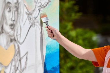 Close-up of female hand painting canvas outdoors, sketching woman face. Natural lighting highlights vivid brush strokes. Green foliage in background clipart