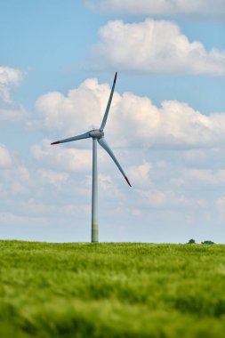 Rüzgar türbini, parlak mavi gökyüzünün altındaki yemyeşil buğday tarlasında kabarık beyaz bulutlarla dimdik ayakta duruyor.