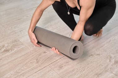 Genç bir kadın, açık ahşap zeminde gri yoga minderi yuvarlıyor. Sahne, yukarıdan bakıldığında minimalist iç mekanlarda sükunet ve hazırlığı çağrıştırıyor.