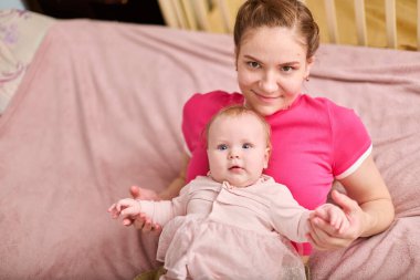 Kahverengi saçlı genç bir kadın pembe elbiseli sevimli bir kızı tutuyor. Avrupa kökenli, her ikisi de kapalı yerlerde yumuşak pembe battaniyede gülümsüyor ve yakınlarda ahşap bir beşik var.
