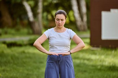 Siyah saçlı genç bir kadın, güneşli parkta qigong çalışıyor. Odaklanmış ifade, silah oluşturma çemberi. Ağaçların ve yumuşak yeşil çimenlerin arkaplanı sükuneti artırır