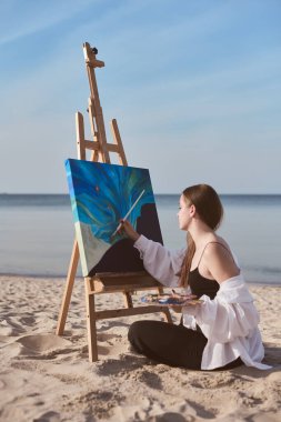 Genç bir kadın kumlu sahilde otururken tuvale soyut bir okyanus manzarası çiziyor. Sakin deniz düzeni yaratıcılığı arttırır, ilham ve huzuru sembolize eder.