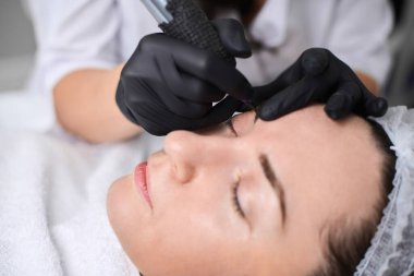 Güzellik uzmanı kuafördeki genç kadın kaşlarına kalıcı makyaj uygular. Odaklanma hassasiyet ve sanatsallık üzerinedir, sakin ortam deneyimi artırır.