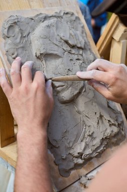 Yetişkin adam ahşap aletler kullanıyor, kil yardım portresi yontuyor. Doğal ışık, dokuları ön plana çıkarır. Ahşap sehpa çerçeveleri çalışma alanı