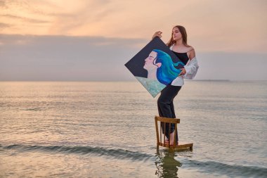 Young female painter stands on chair in serene water, holding vibrant portrait. Peaceful seaside setting at sunset reflects creativity and tranquility clipart