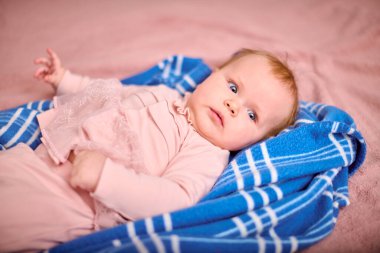 Kızıl saçlı ve açık tenli bir bebek, mavi çizgili battaniyenin üzerinde yatıyor. Yumuşak ışıklandırma pembe elbiseyi aydınlatır, huzur ve rahat ortam yaratır.