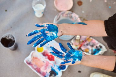 Açık havada serpiştirilmiş sanat malzemeleriyle dolu canlı mavi boyayla kaplı yetişkin elleri. Yumuşak ışıklandırma yere yumuşak gölgeler düşürür, sanatsal havayı geliştirir.