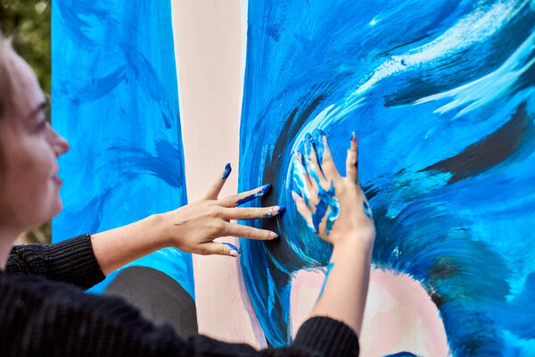 Молодая женщина со светлой кожей использует пальцы, чтобы рисовать на холсте. Яркие голубые вихри доминируют в произведениях искусства, установленных в пышном саду при мягком дневном свете