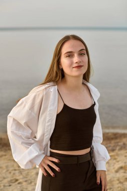 Genç bayan sakin deniz kenarında güven içinde duruyor. Onun rahat duruşu ve nazik gülümsemesi, sakin ve huzurlu titreşimler iletir. Gün batımının yumuşak parıltısı rahatlatıcı atmosferi canlandırır.