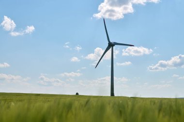 Rüzgar türbini, parlak mavi gökyüzünün altındaki yemyeşil buğday tarlasında kabarık beyaz bulutlarla dimdik ayakta duruyor.