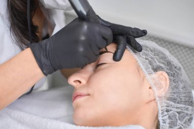 Genç kadın, temiz ve steril bir şekilde kaş makyajına maruz kalıyor. Eldiven giyen profesyonel teknisyen, hassas ve hijyen sağlayan pigmentleri dikkatlice uyguluyor.