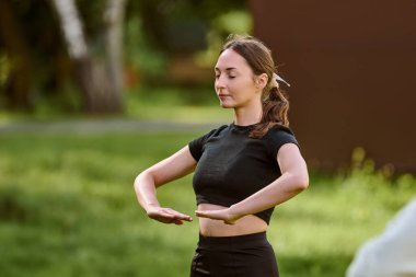 Genç beyaz kadın güneşli parkta qigong çalışıyor. Kahverengi saçlı, arkadan bağlanmış, siyah elbiseli, sakin yüzlü. Yeşil çevre, yumuşak doğal ışık