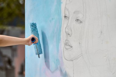 El, beyaz duvardaki genç kadın yüzünün çiziminin üzerine yumuşak mavi boya sürüyor. Doğal dış mekan, hafif ışıklandırma, sanatsal gelişim