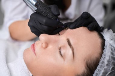 Güzellik uzmanı kuafördeki genç kadın kaşlarına kalıcı makyaj uygular. Odaklanma hassasiyet ve sanatsallık üzerinedir, sakin ortam deneyimi artırır.