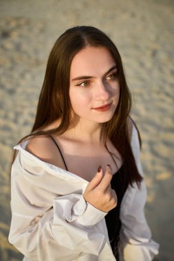 Genç bir kadın kumlu sahilde duruyor, gevşek, açık beyaz bir gömlek giyiyor. Onun sakin ifadesi ve yumuşak ışığı huzurlu ve huzurlu bir ruh hali uyandırıyor, yansıma anını işaret ediyor.