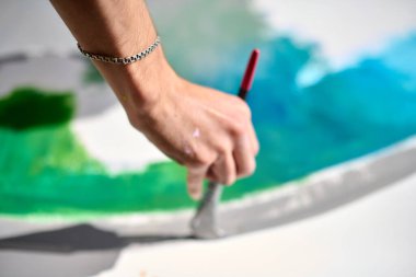 Genç, açık tenli, erkek sanatçı tuvale renkli ve soyut bir eser çiziyor. Mavi ve yeşil, yumuşak ışıklandırma ve odaklanmış perspektifle baskın sahne