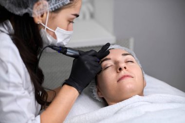 Genç bir kadın huzurlu bir kaplıcada kaş makyajı yaptırıyor. Güzellik teknisyeni, eldiven takıyor, dikkatle odaklanıyor, sakin, profesyonel atmosfer yaratıyor.