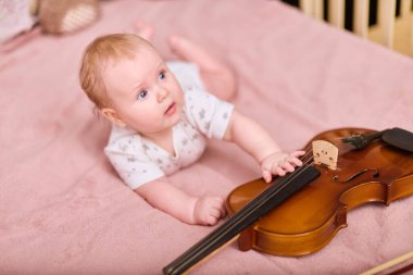 Sarı saçlı küçük kız pembe yatağa uzanmış, garip bir şekilde kemana uzanıyor. Konforlu yatak odası ayarları, sıcak ışıklandırma huzurlu ve neşeli atmosferi geliştirir.