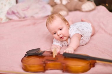 Sarışın, beyaz bir kız bebek, yatağa uzanıp keman izliyor. Yumuşak ışıklandırma, sıcak ortam. Pembe yatak örtüsü daha yumuşak bir zemin ekler, merakına odaklan.