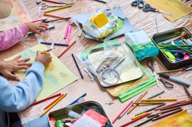Çocuklar resim malzemeleriyle dolu bir masada resim yaparlar. Parlak renkler, yoğun ortam ve yukarıdan aşağı perspektif enerjik bir atmosfer yaratır