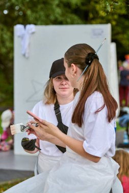 Young adult caucasian women painting outdoors at festival. Intense focus, creative atmosphere, brushes in hand. Background features greenery and easel clipart