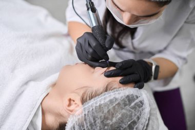 Yetenekli teknisyen, sakin salondaki genç bayana kalıcı kaş makyajı yapıyor. Siyah eldivenlerle, profesyonel aletler kullanılıyor, hassas ve rahatlatıcı ortamlarda bakım sağlanıyor.