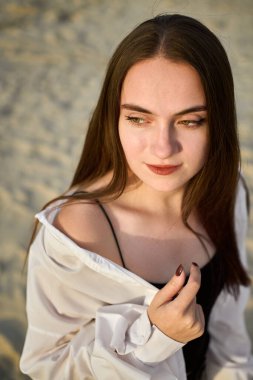 Genç bir kadın kumlu sahilde duruyor, gevşek, açık beyaz bir gömlek giyiyor. Onun sakin ifadesi ve yumuşak ışığı huzurlu ve huzurlu bir ruh hali uyandırıyor, yansıma anını işaret ediyor.