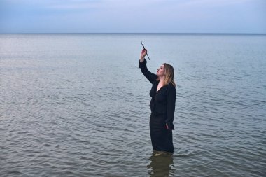 Young woman in black dress stands waist-deep in calm sea waters, holding paintbrush. Tranquil seascape suggests themes of magic and contemplation under soft, blue sky clipart