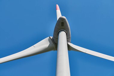 Güneşli bir günde açık mavi gökyüzüne karşı açılan kırmızı uçlu rüzgar türbininin yakın görüntüsü, berraklığı ve çevresel sürdürülebilirliğe bağlılığı çağrıştırıyor.