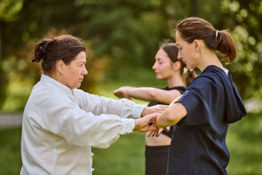 Üç kadın, yetişkin ve genç, yemyeşil bahçede qigong oynuyorlar. Sabah ışığı odaklanmayı arttırır. Beyaz ve koyu renk elbiseler canlı yeşilliklerle çelişiyor.
