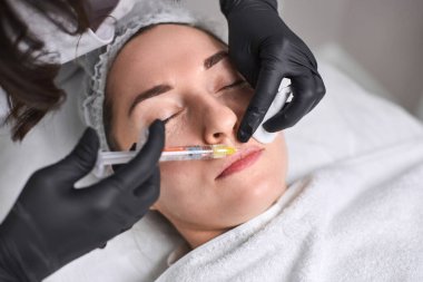 Profesyonel yöneticiler, klinik ortamdaki cerrahi olmayan dudak geliştirmelerinde hassasiyet ve bakımı vurgulayan, gevşemiş genç kadına deri doldurucu enjeksiyonu yapar.