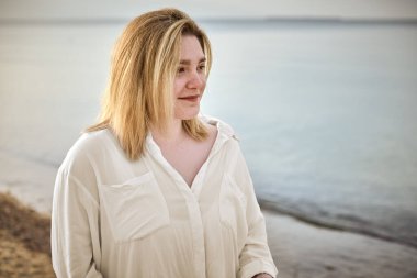 Beyaz bluzlu genç bir kadın sakin kumsalda duruyor, sakin denize özenle bakıyor, huzur ve huzur hissi uyandırıyor.