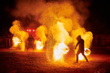 Gece gökyüzünde parıldayan iki itfaiyeci silueti, bir yangın gösterisi sırasında enerji ve ateş manipülasyonu gösteriyorlar.
