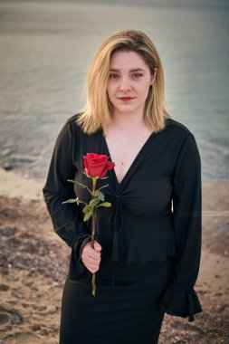 Siyahlı genç bir kadın, elinde kırmızı gülle sakin bir plajda duruyor. Sakin deniz ve yumuşak ışık, aşkı ve hatıraları sembolize eden, yansıtıcı havayı çağrıştırır.