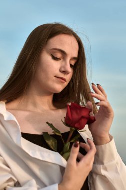 Beyaz gömlekli genç bir kadın nazikçe kırmızı bir gül tutuyor, sükunet saçıyor. Huzur veren gökyüzü, doğanın ortasında güzelliği ve içselliği sembolize eder.