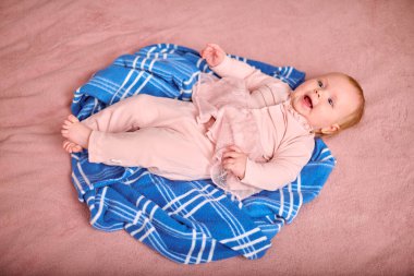 Mavi kareli battaniyeye uzanmış açık renk saçlı genç bir kız çocuğu. Pembe giyinmiş, neşeli görünüyor. Yumuşak pembe tonlarda arkaplan, yukarıdan çekilen