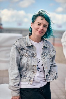 Açık havada kot ceketli, kısa mavi saçlı genç beyaz kadın. Yumuşak bir gülümseme, rahat bir poz, bulutlu bir arka plan, rüzgarlı bir gün, doğal ışık.