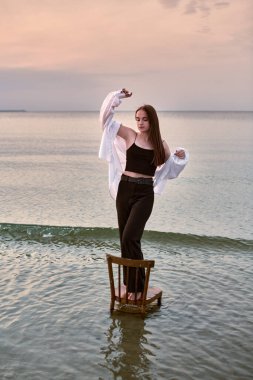 Genç bir kadın gün batımında sahildeki sandalyede güven içinde duruyor. Sakin su ve yumuşak parıltı denge ve huzuru sembolize eden barışçıl bir ortam yaratır.