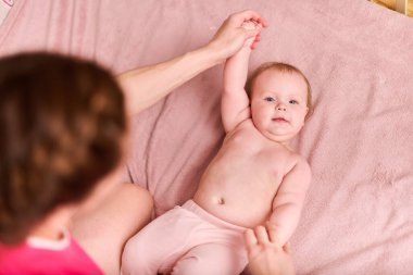 Genç bir anne, bebeğe pasif egzersizlerde yardım ediyor. Pembe battaniyeye uzanmış bebek, rahat bir ortam, sıcak bir ışık. Kadın beslenme üzerine odaklanır