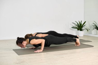 Siyah aktif giysili iki genç kadın parlak stüdyodaki yoga minderlerinde poz veriyorlar. Yerler hafif ahşap zeminler ve yeşil bitkilerle sükunetli bir atmosfer yaratıyor.