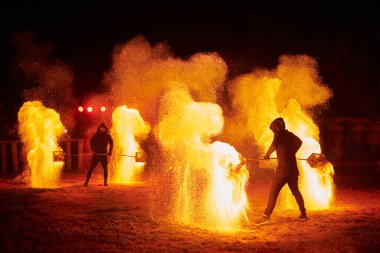 Gece gökyüzünde parıldayan iki itfaiyeci silueti, bir yangın gösterisi sırasında enerji ve ateş manipülasyonu gösteriyorlar.