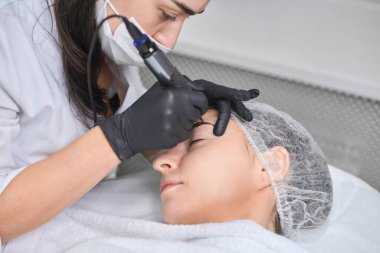 Genç kadın, temiz ve steril bir şekilde kaş makyajına maruz kalıyor. Eldiven giyen profesyonel teknisyen, hassas ve hijyen sağlayan pigmentleri dikkatlice uyguluyor.